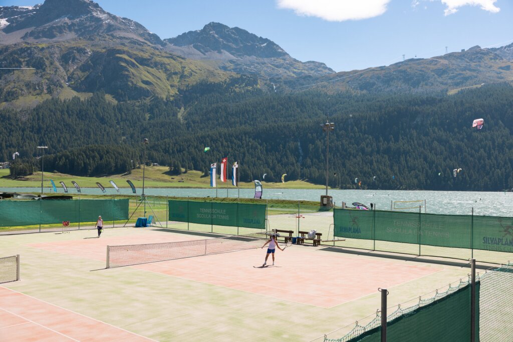 Two people are playing tennis on a court by Lake Silvaplana, with forests and the Corvatsch mountain in the background.